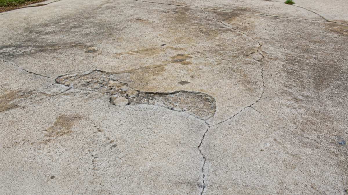 Close-up of concrete driveway surface showing visible cracks, spalling, and discolored patches that indicate damage requiring repair before resurfacing